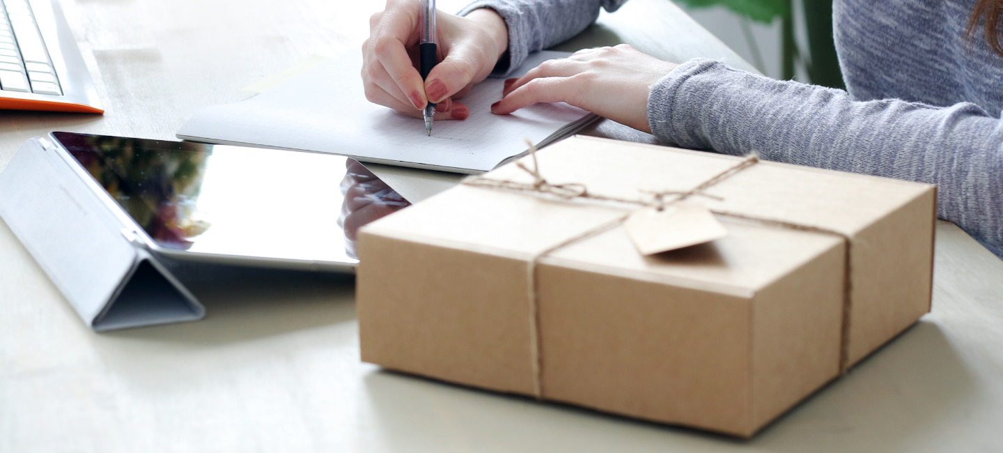 A imagem mostra uma pessoa a escrever num papel, com uma caixa de cartão em primeiro plano e um portátil ao fundo.