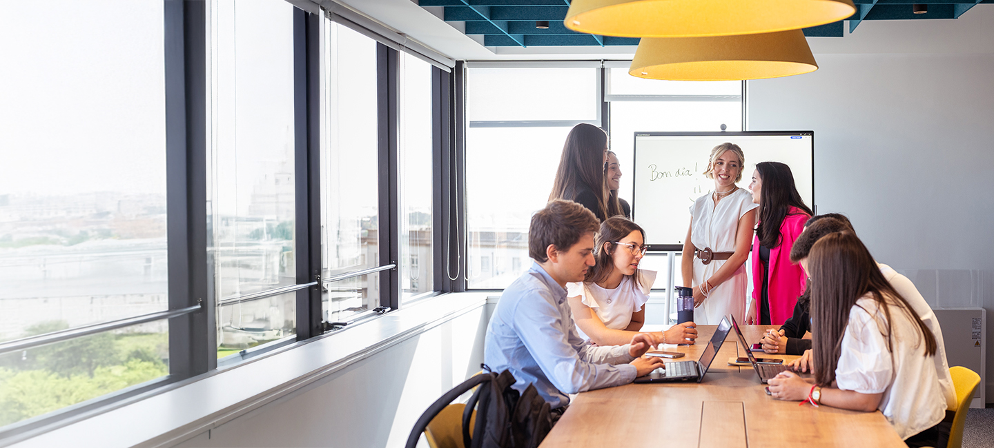 Mesa com 4 jovens sentados  com computadores e dois a apresentar num quadro