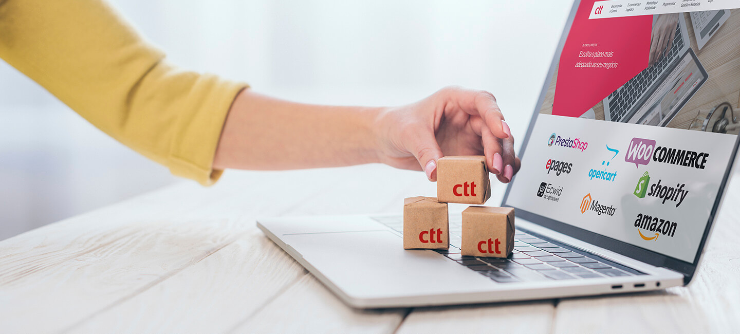 Pessoa a colocar pequenas caixas do CTT sobre o teclado de um portátil com logótipos de plataformas de e-commerce no ecrã.