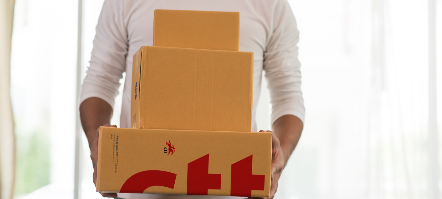 Pessoa segura três caixas de cartão empilhadas, a caixa maior tem o logótipo dos CTT em vermelho.