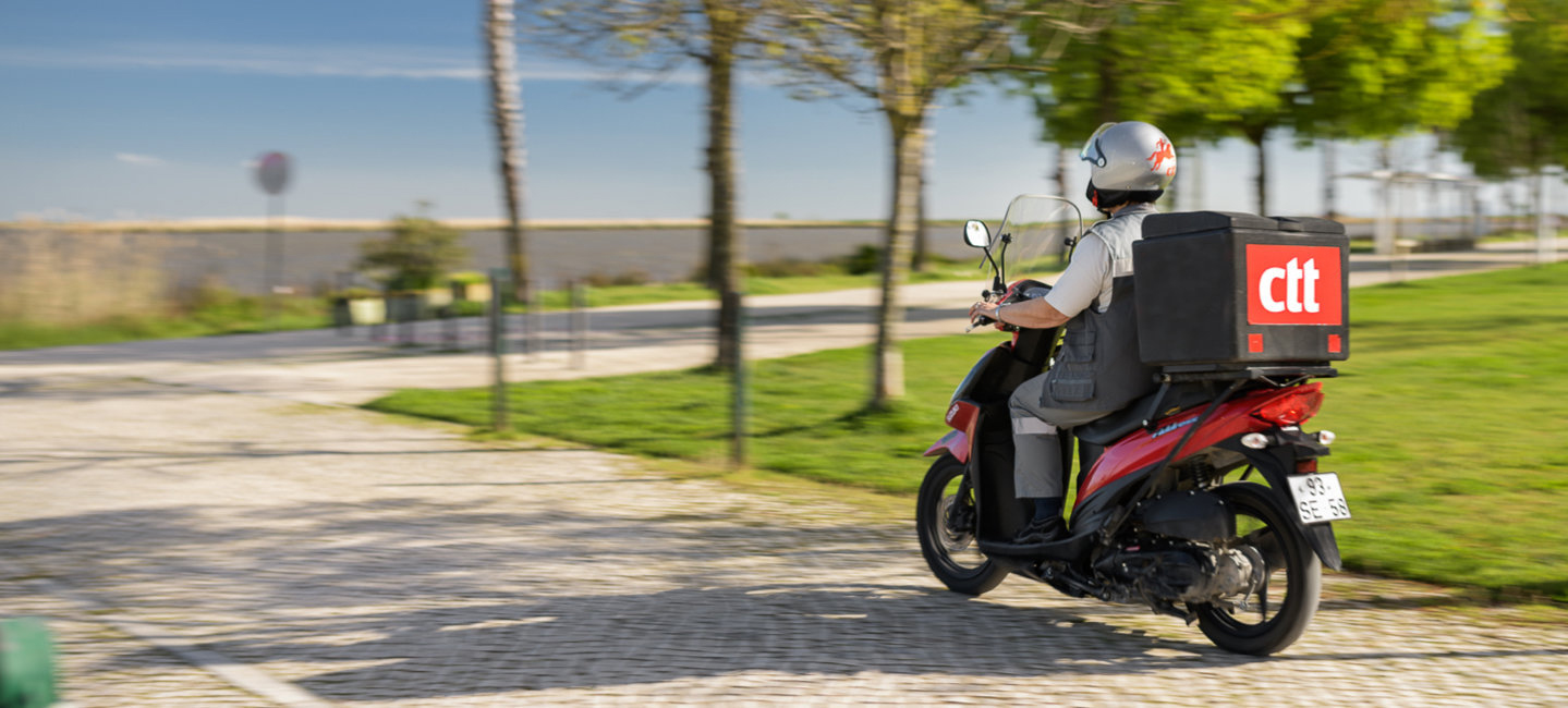Pessoa a conduzir uma mota vermelha com o logótipo dos CTT, para distribuir correio.