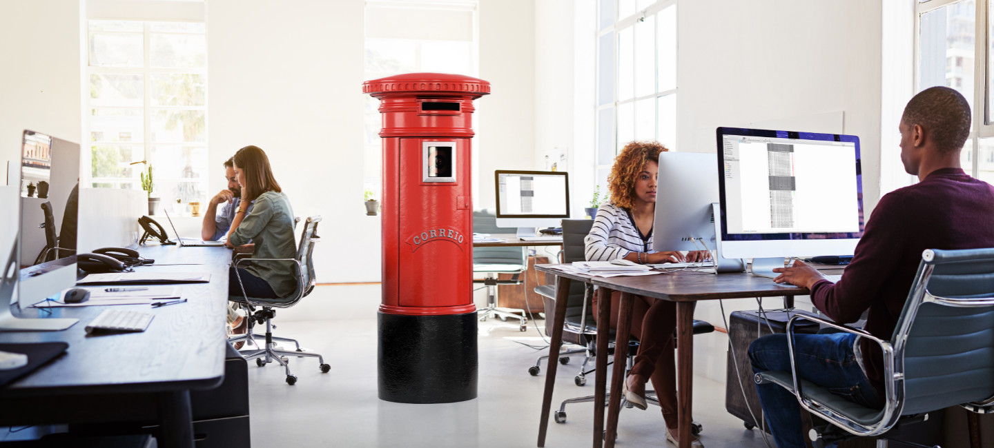 Ambiente de escritório com pessoas a trabalhar e um marco de correio vermelho no centro da sala.